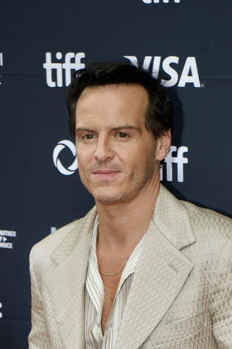 Andrew Scott in a cream suit and open shirt on a festival carpet, hands loosely clasped
