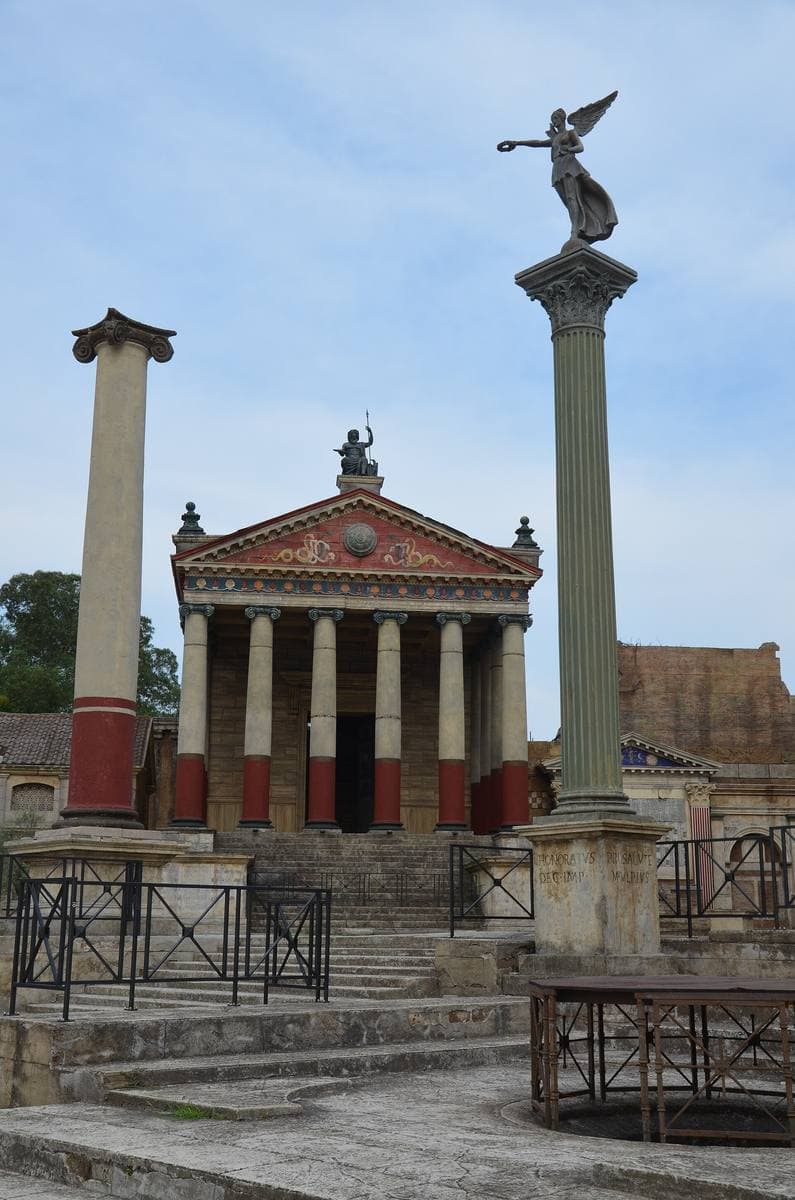 A tall column topped by a winged figure in front of a red-columned temple