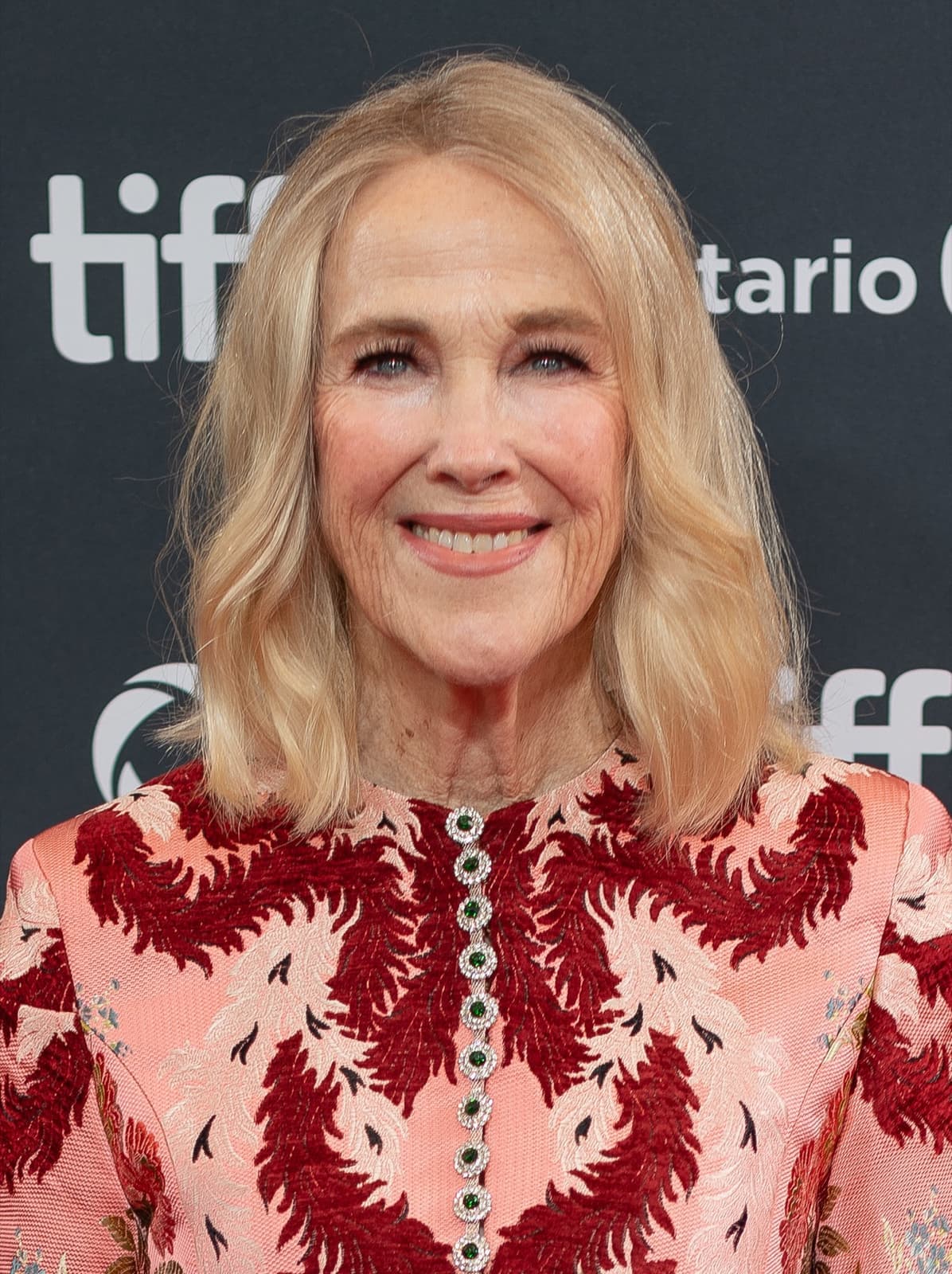Catherine O’Hara smiling, shoulder-length blonde hair, in a pink and red patterned dress