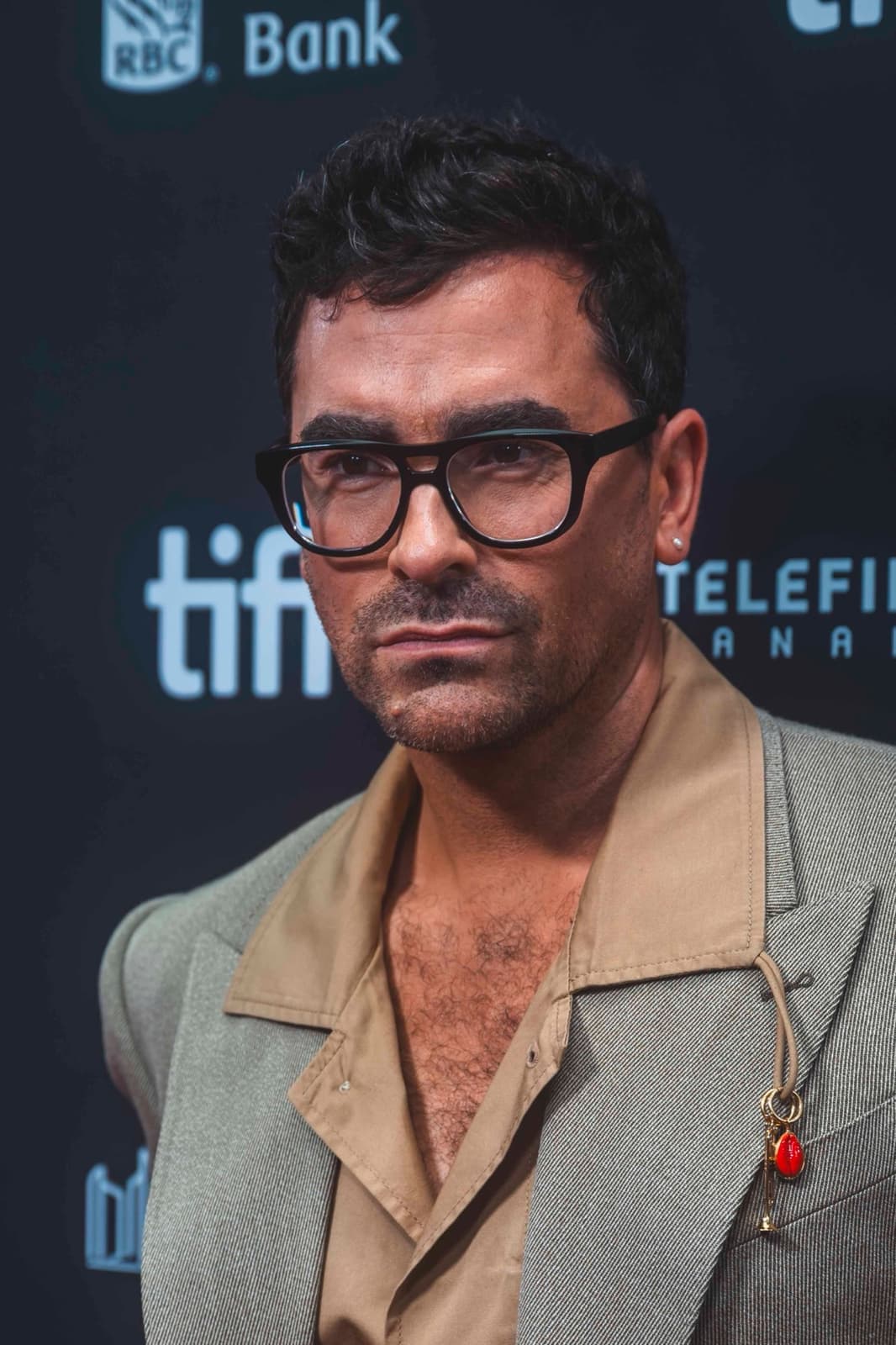 Dan Levy in heavy black glasses and a grey jacket over a camel shirt