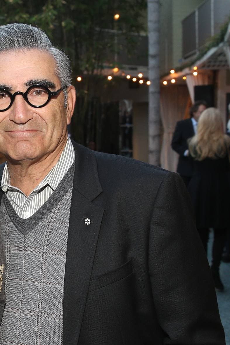 Eugene Levy in round black glasses, a grey sweater vest and dark jacket, smiling