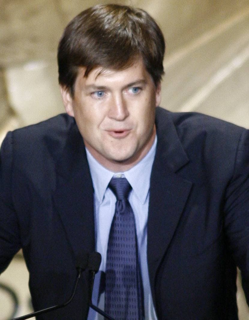 A man in a dark suit and blue tie speaking at a lectern microphone