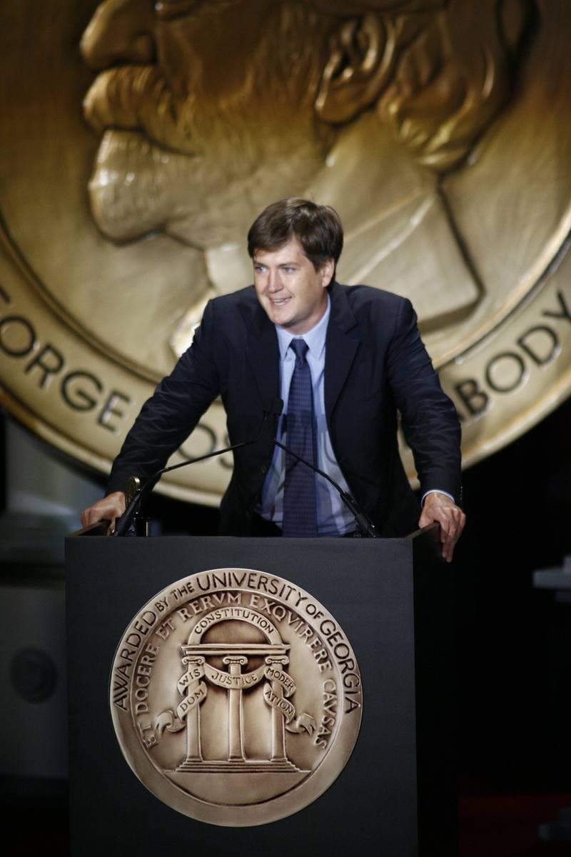 A man in a suit at a lectern in front of a giant gold medal