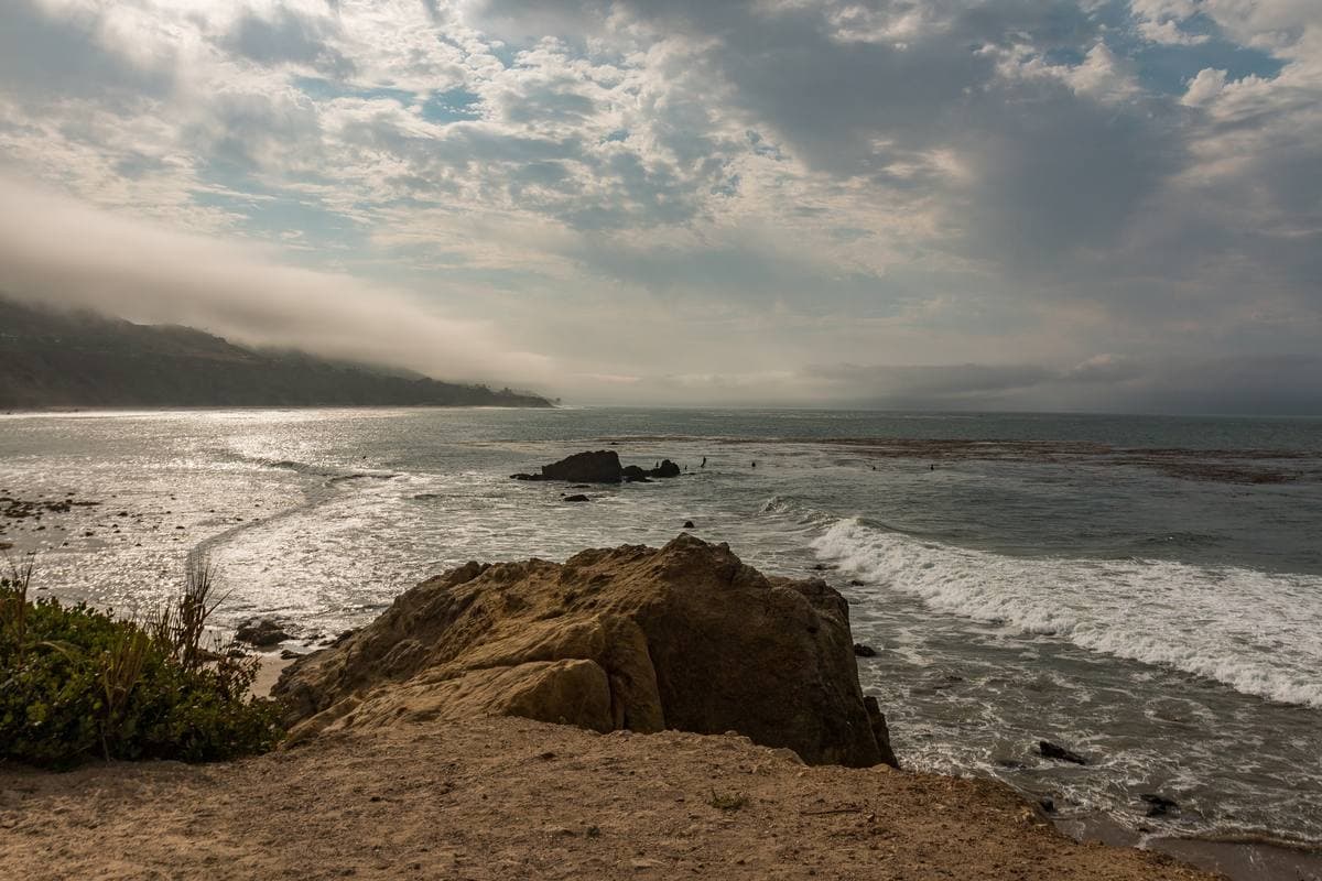 Waves breaking on a rocky point under low cloud, with a hazy headland behind