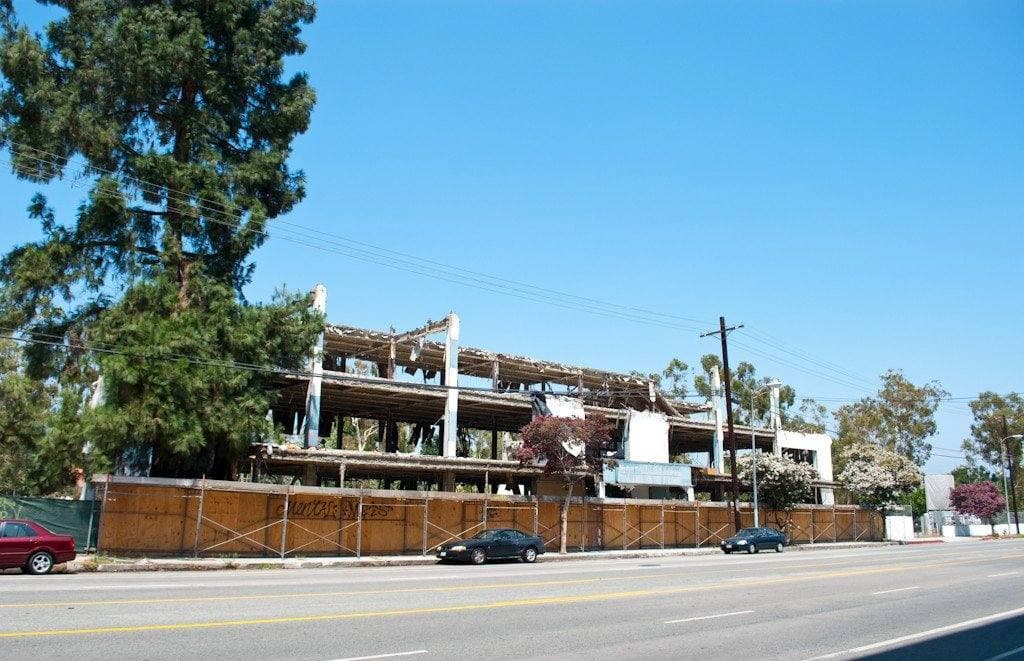 A low concrete building stripped to its floors and columns behind a construction fence on a wide sunny boulevard