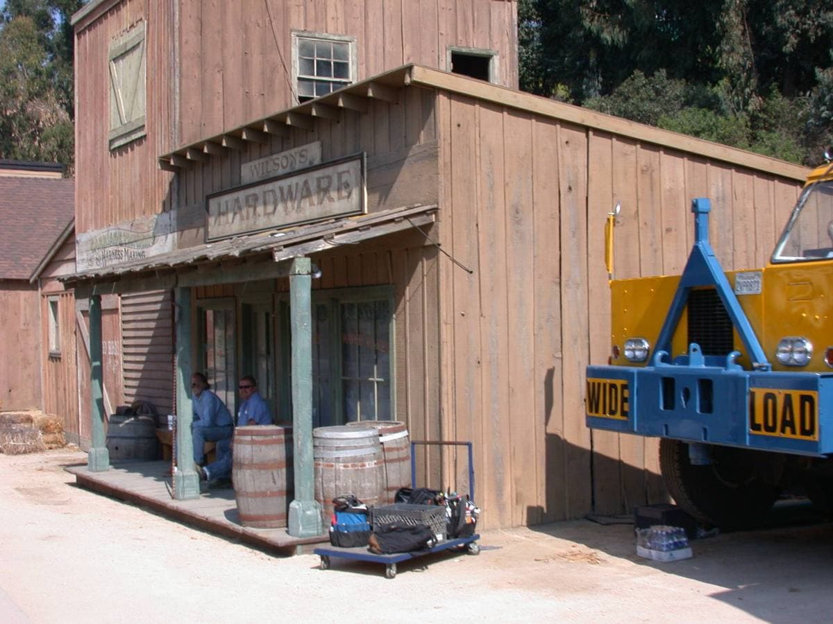 A wooden western-town street set with a timber storefront, barrels and two workers resting in the shade