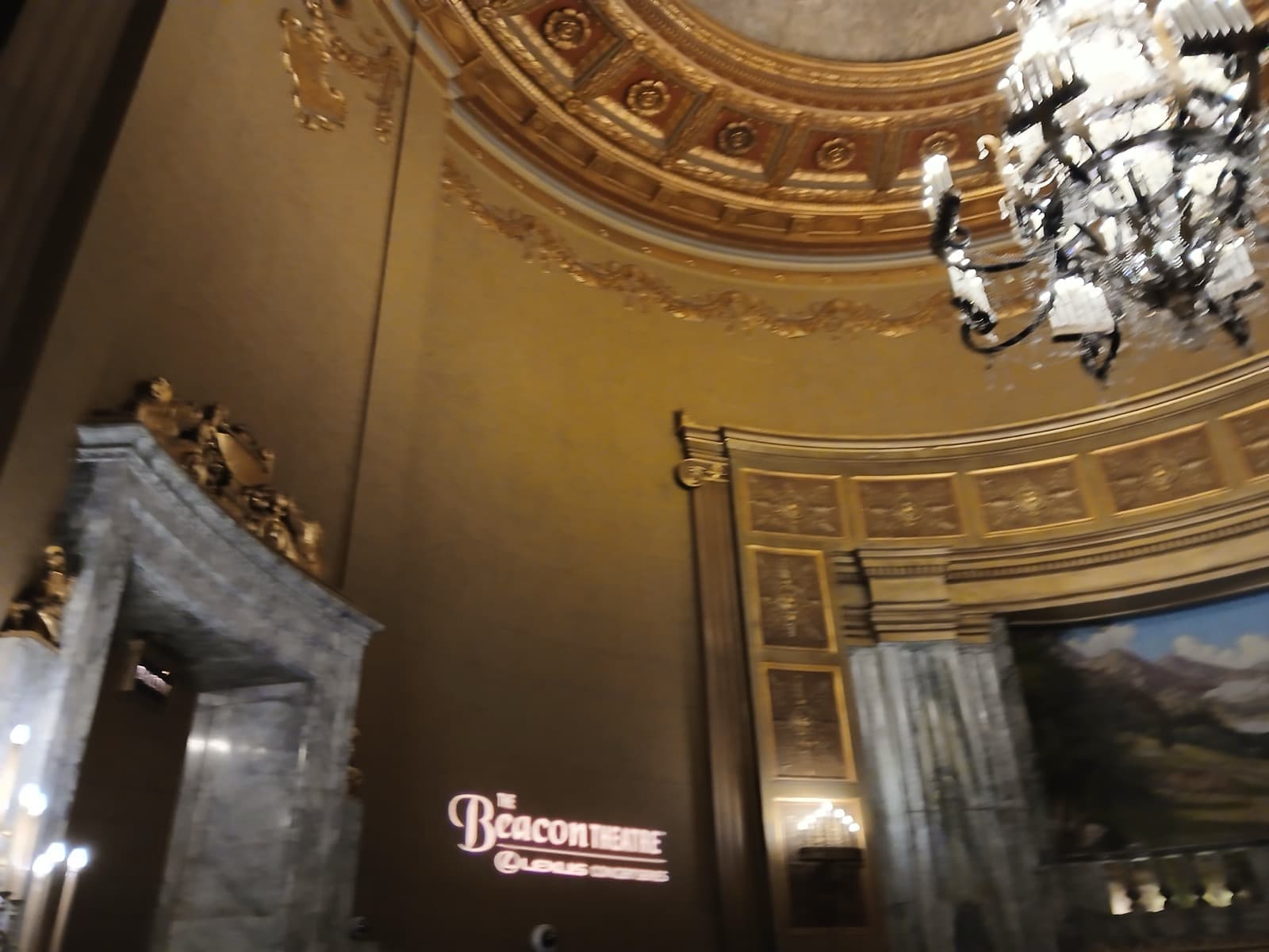 The gilded rotunda of the Beacon Theatre lobby with a chandelier and the theatre name lit on the wall