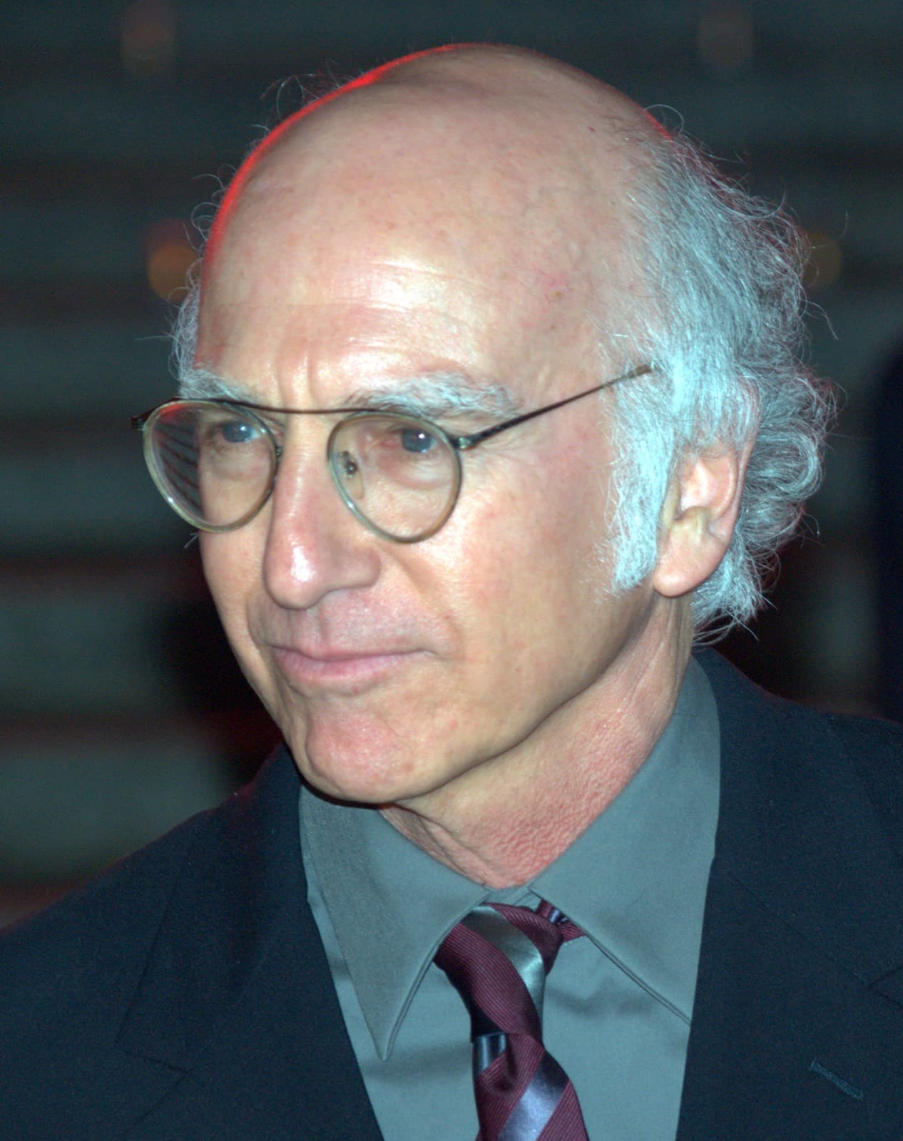 Larry David in round glasses and a dark jacket, looking down to one side