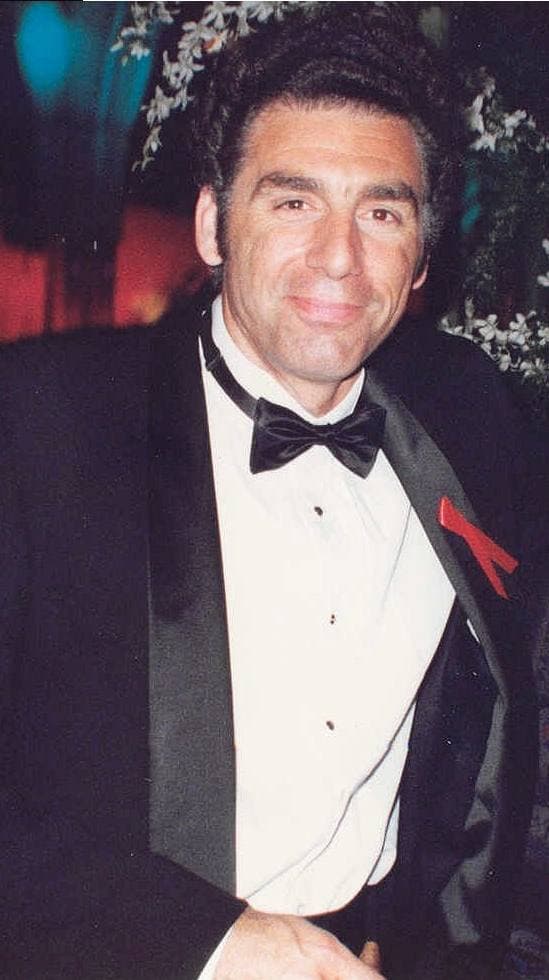 Michael Richards in a tuxedo and bow tie with a red ribbon