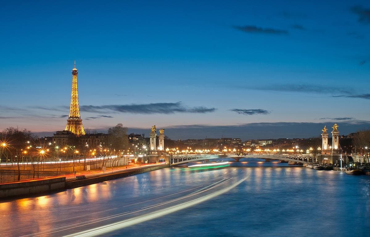 The lit Eiffel Tower across the river with a bridge in the foreground