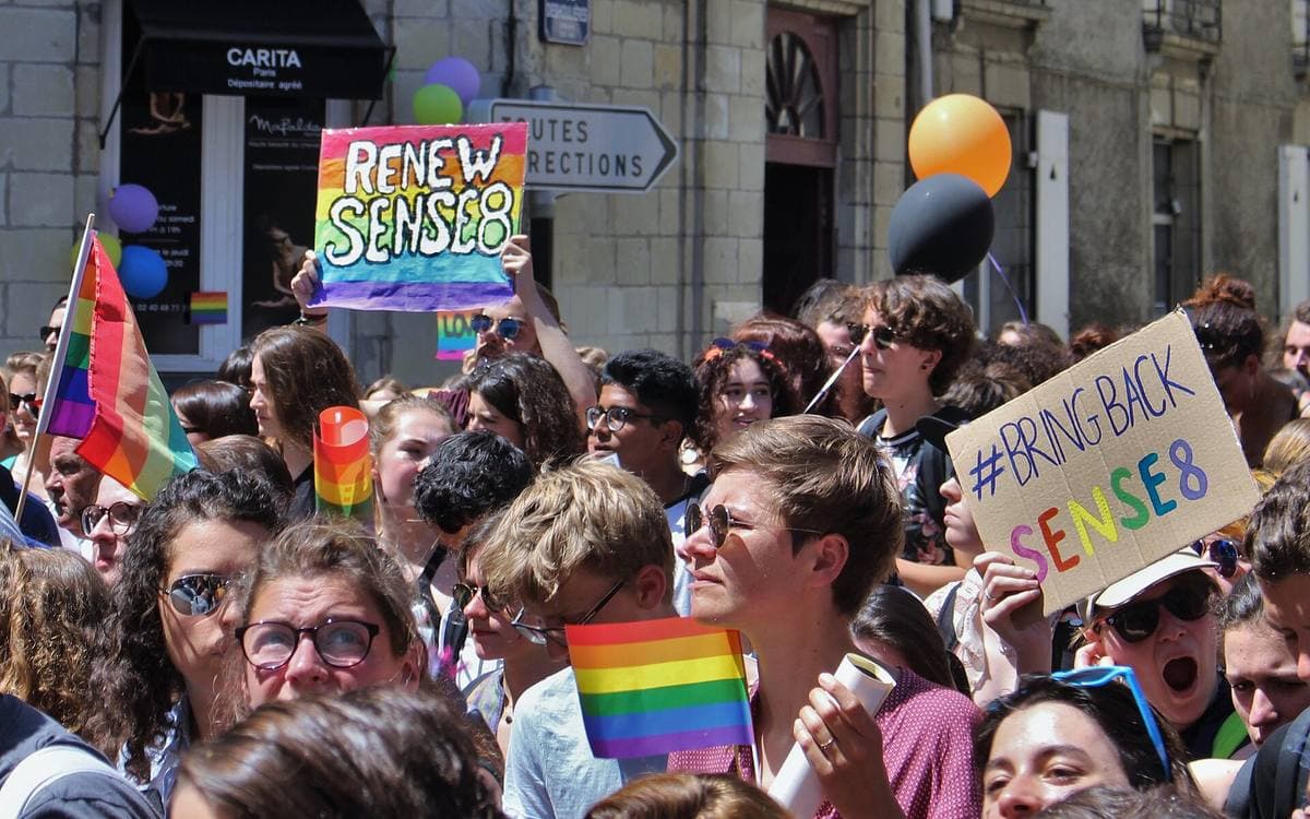 A crowd at a Pride march holding rainbow flags and hand-lettered signs asking for Sense8 to be renewed