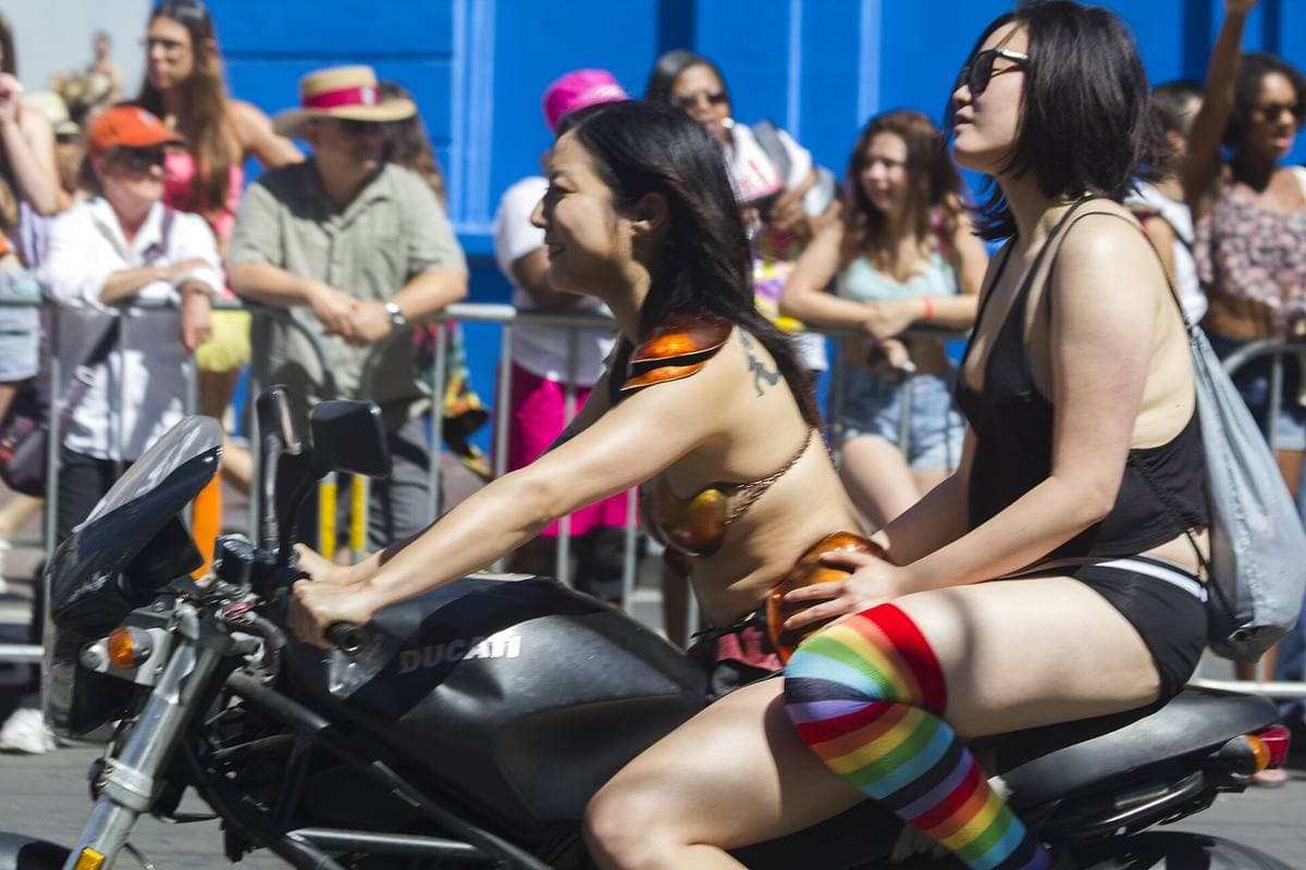Two women on motorcycles in a parade, one in rainbow knee socks, watched by a crowd behind barriers