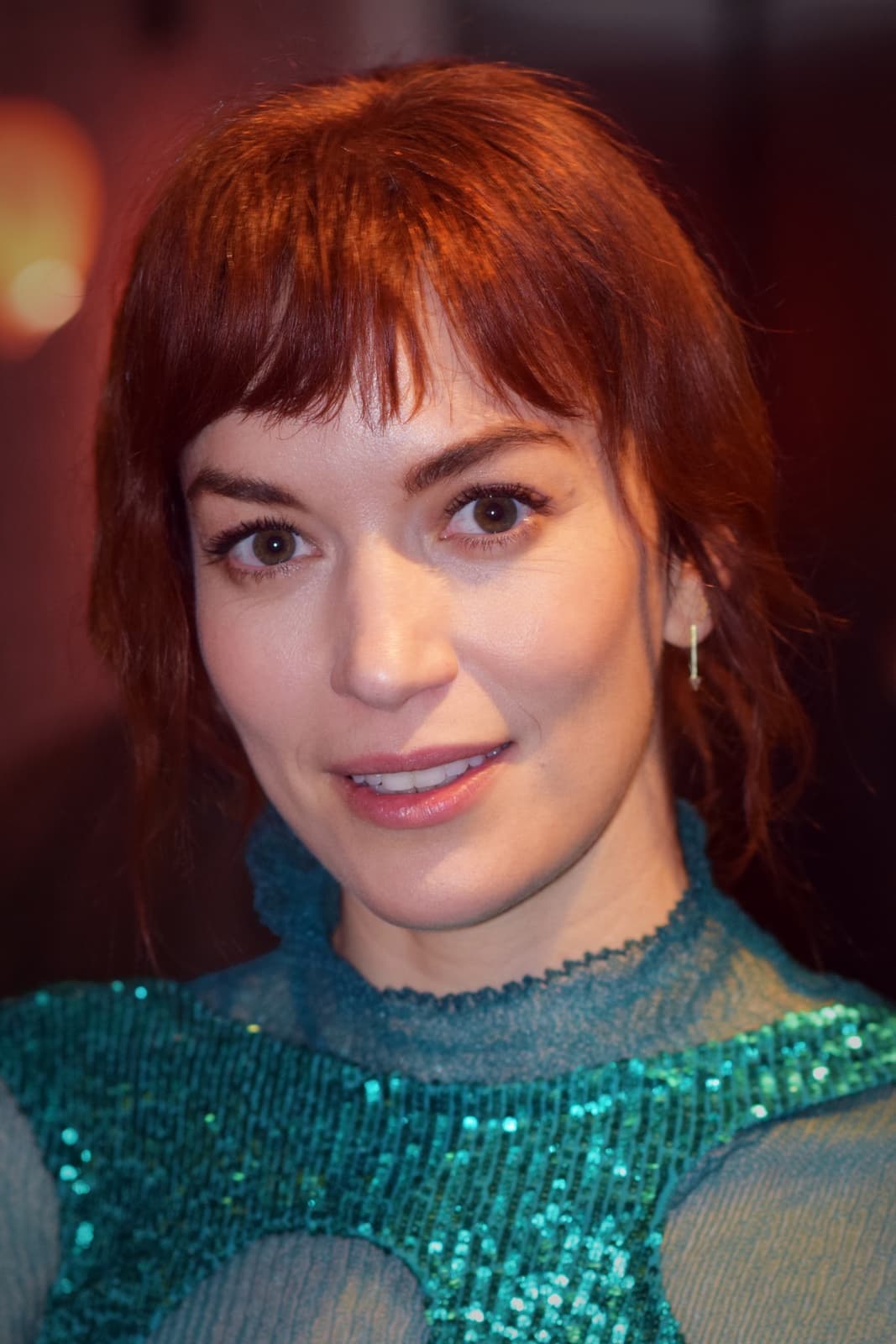Britt Lower with red hair and a fringe, smiling, in a green sequinned top