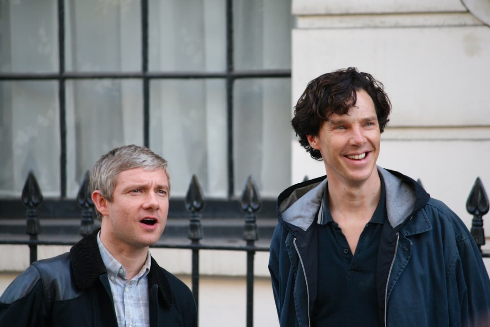 Martin Freeman and Benedict Cumberbatch, smiling, in front of black railings and a pale London façade