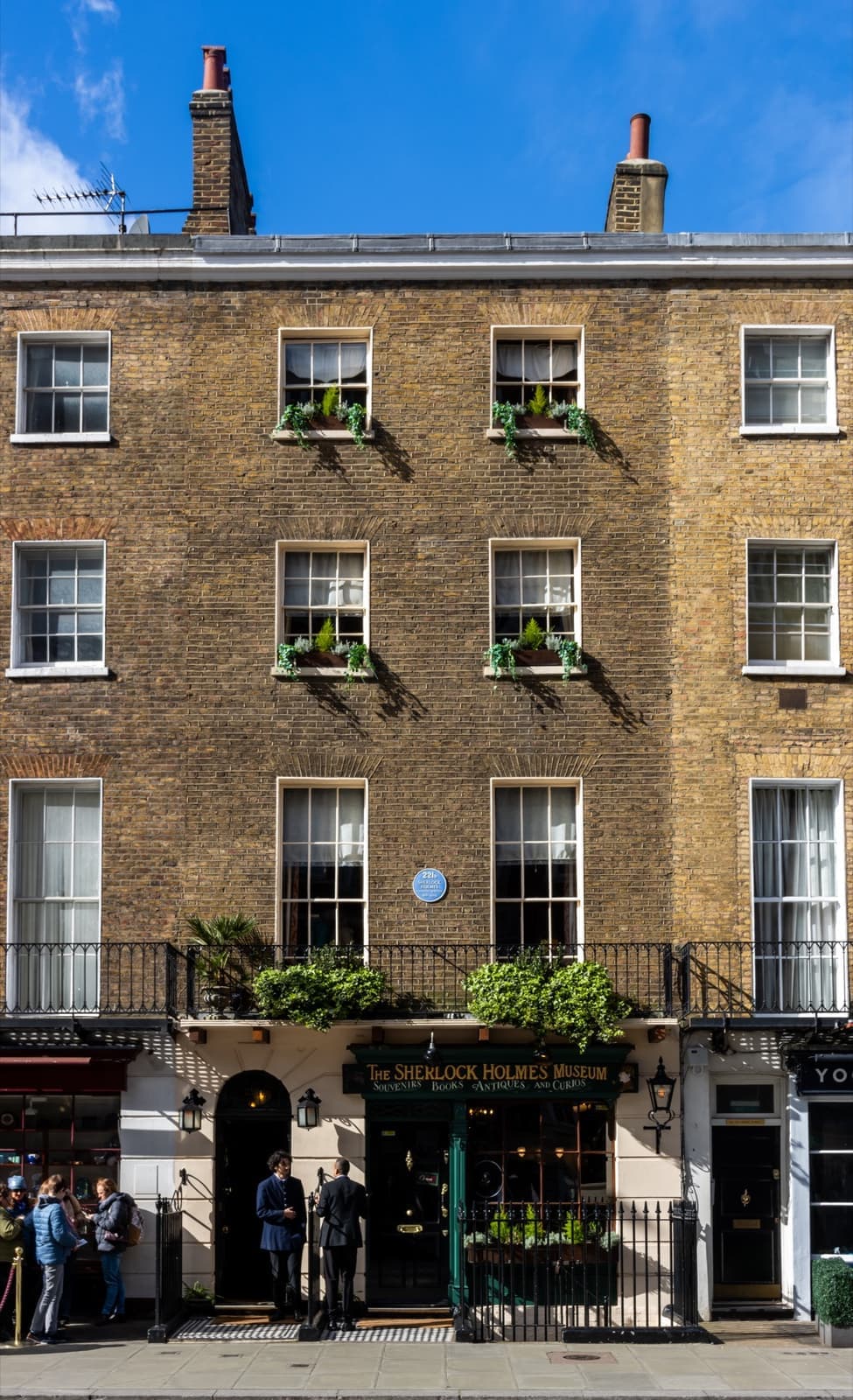A Georgian brick house on Baker Street with a green shopfront and a queue at the door
