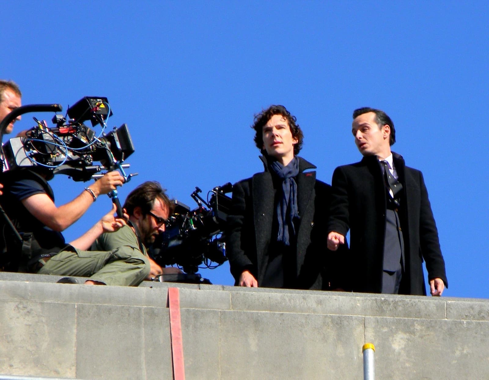 Two actors in dark coats stand on a stone parapet against a blue sky while camera operators film them