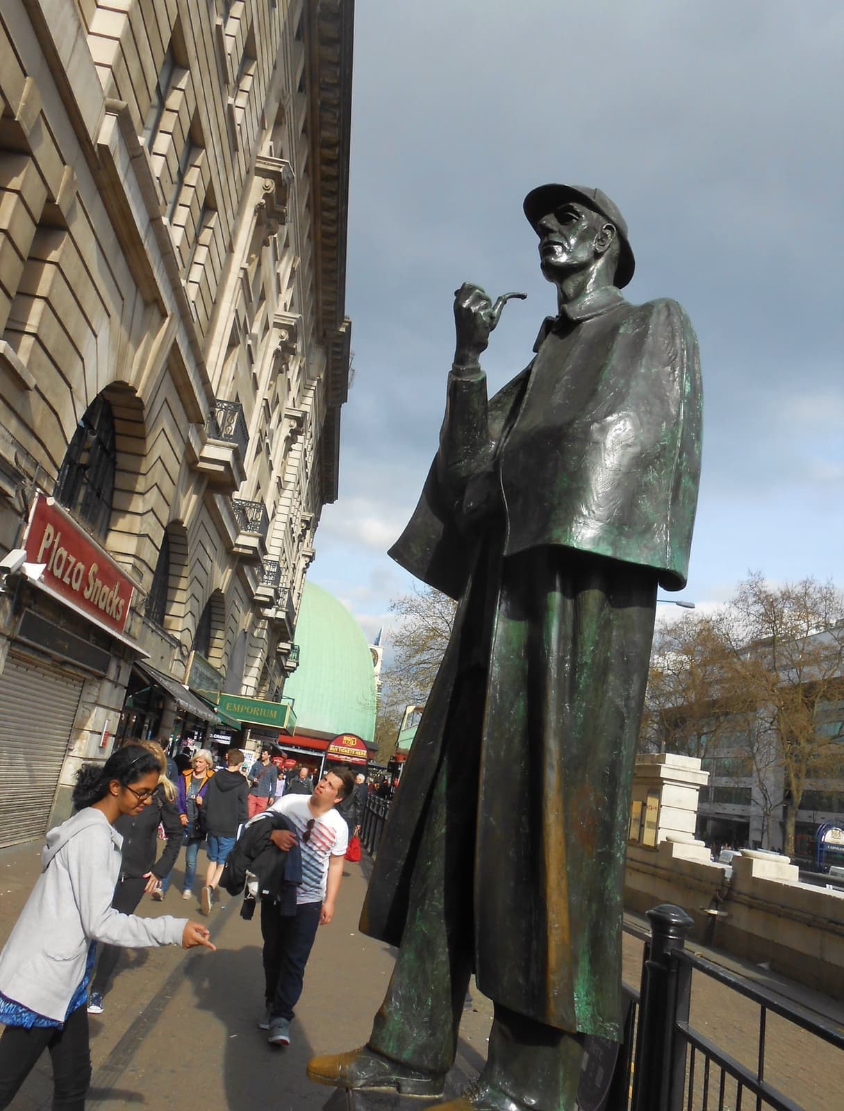 A bronze Sherlock Holmes in deerstalker and cape, holding a pipe, beside a busy pavement