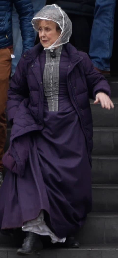 Una Stubbs in a purple Victorian dress and a rain hood, stepping down stone stairs