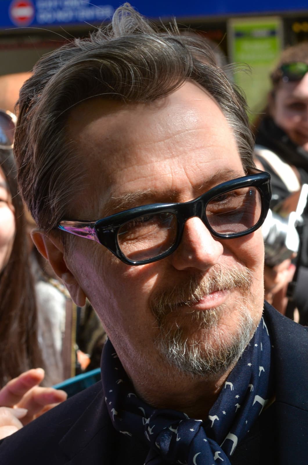Gary Oldman in heavy black glasses and a patterned scarf, half-smiling, in a crowd