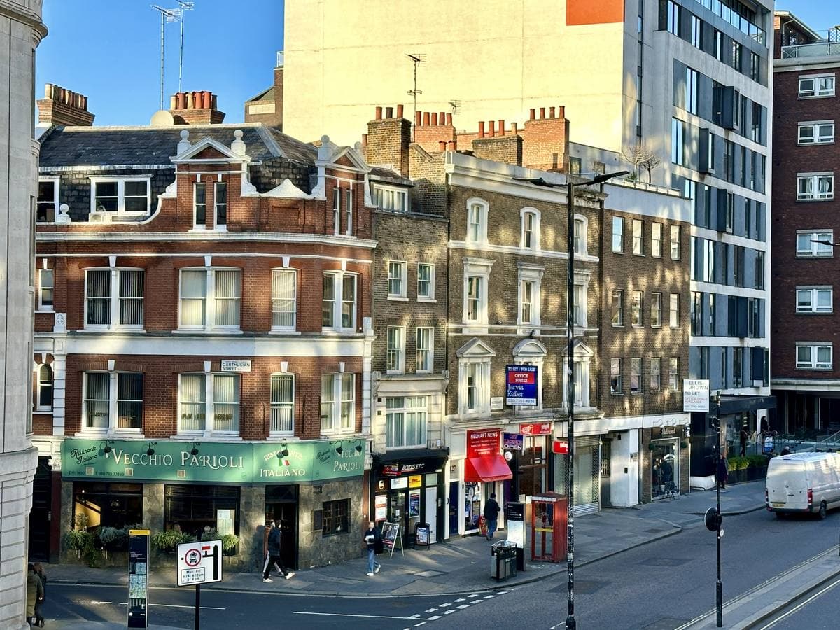 A red-brick Victorian corner building with a green Italian restaurant fascia, a taller pale office block behind, on a sunny City street