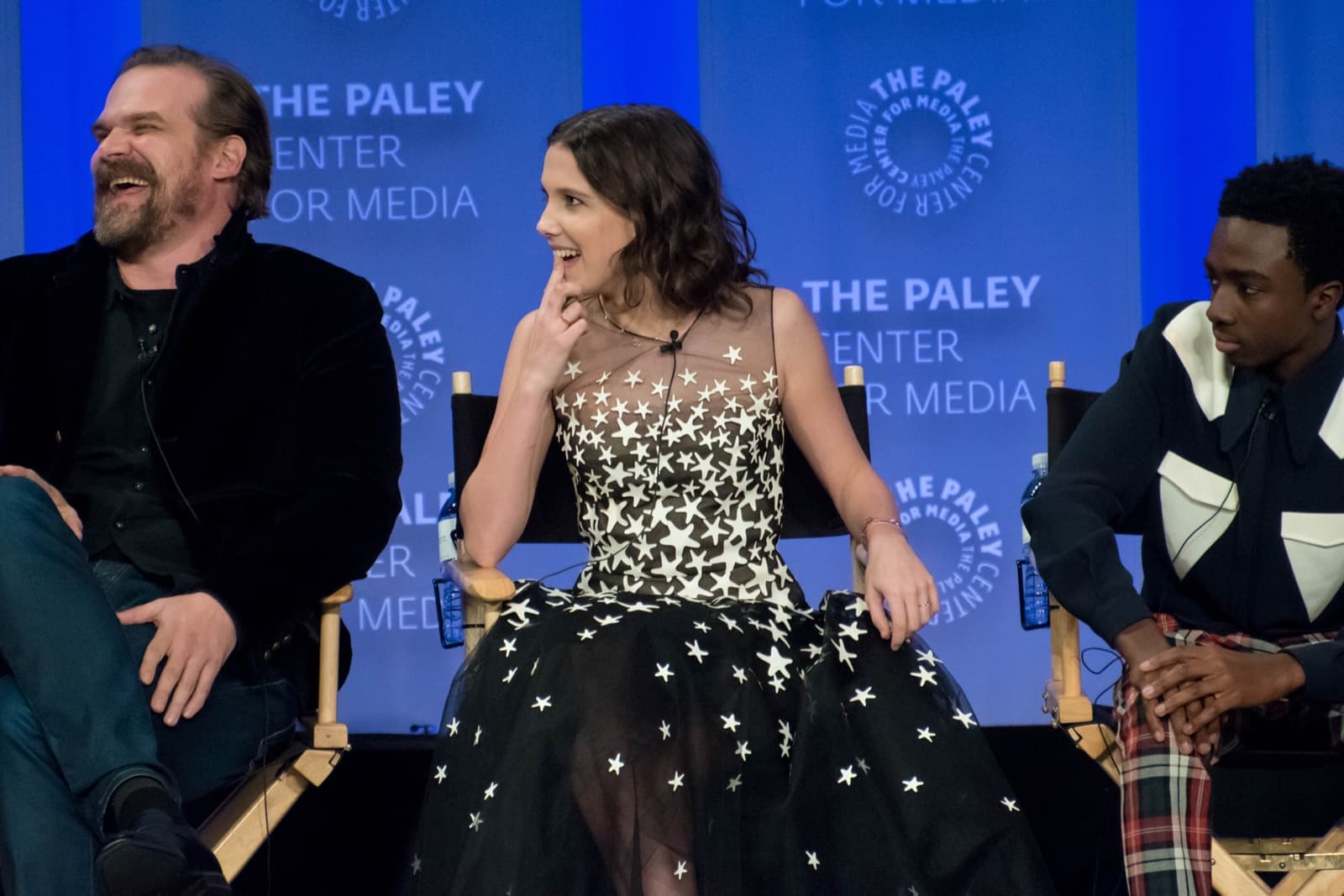 David Harbour laughing beside Millie Bobby Brown and Caleb McLaughlin on a stage in front of a blue backdrop