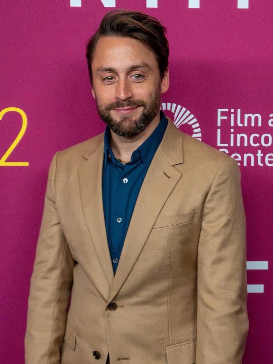 Kieran Culkin in a camel suit and dark teal shirt on a magenta backdrop, half-smiling