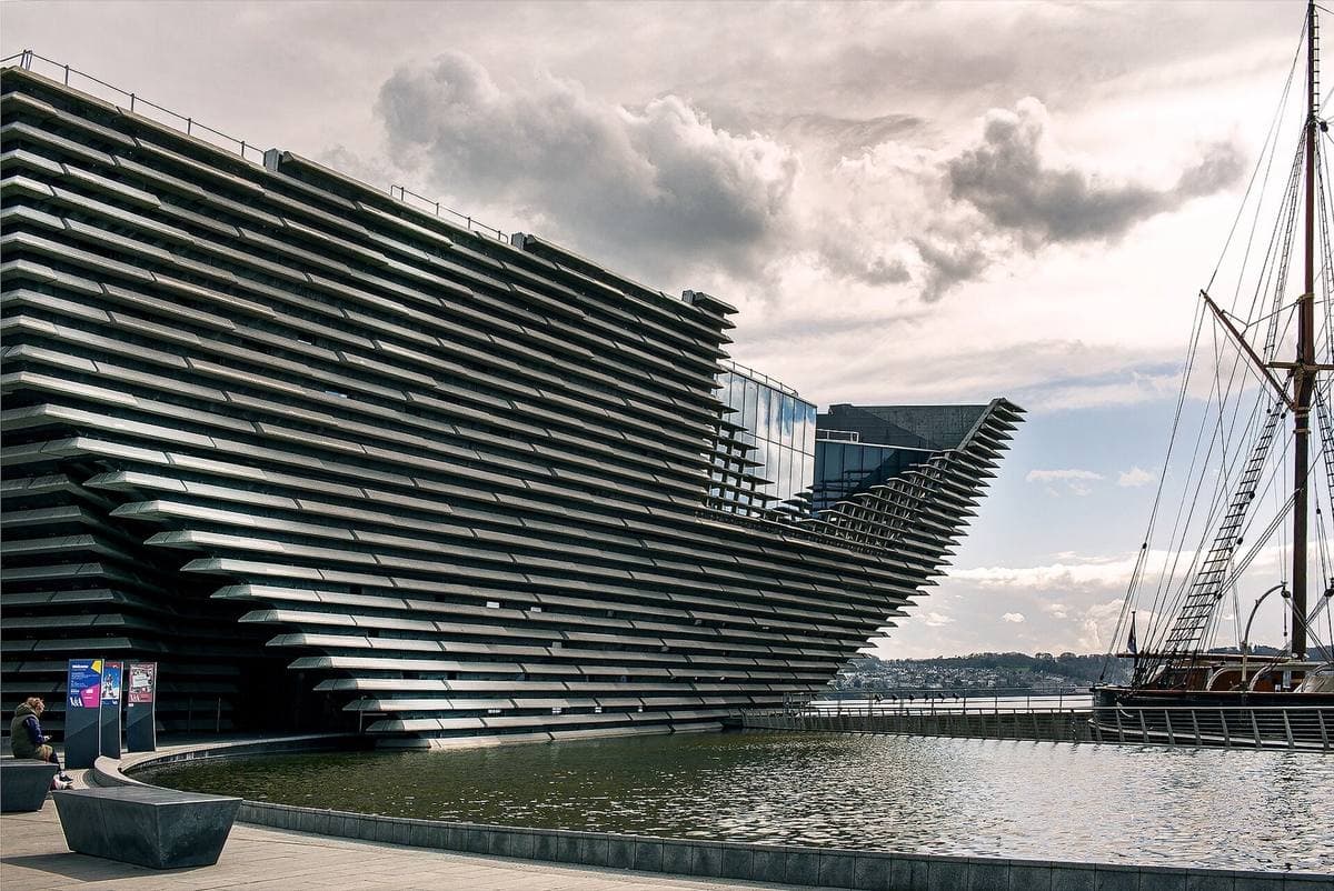 The ribbed, ship-like concrete prow of V&A Dundee over water, with a tall ship moored alongside