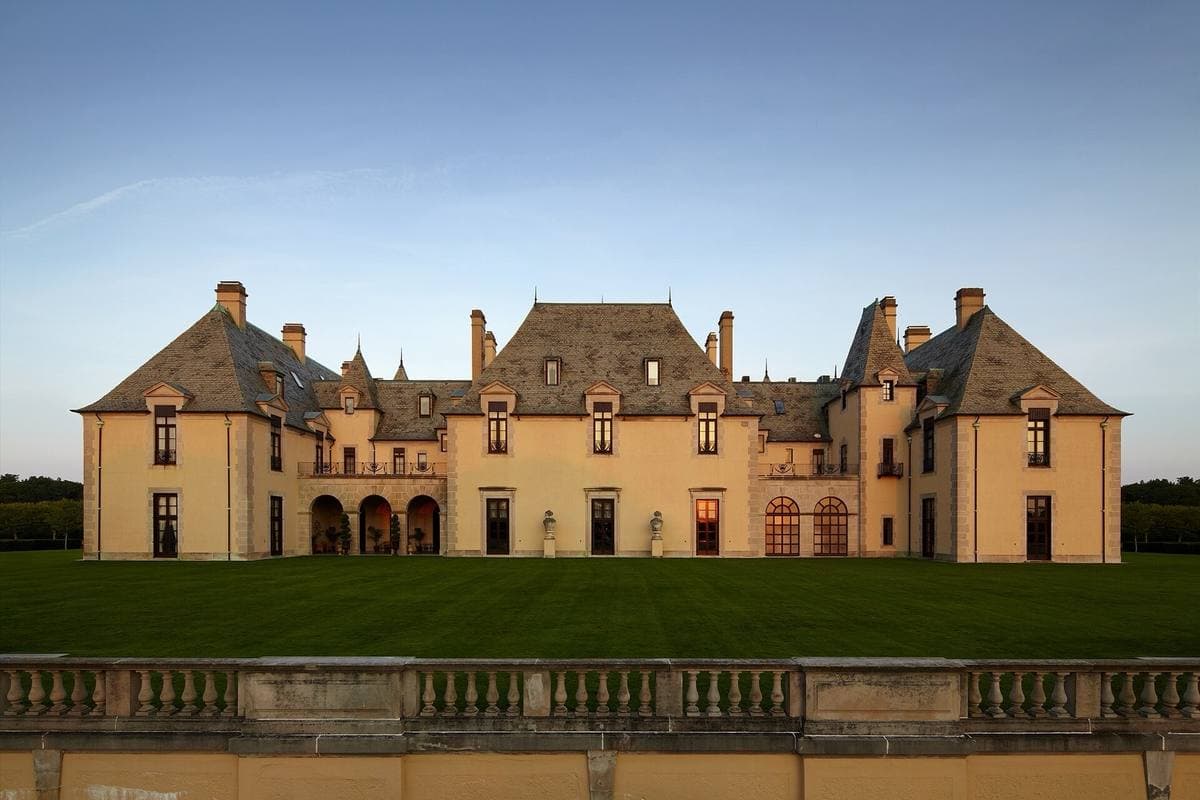 A French-château-style mansion with steep slate roofs in evening sun across a lawn and balustrade