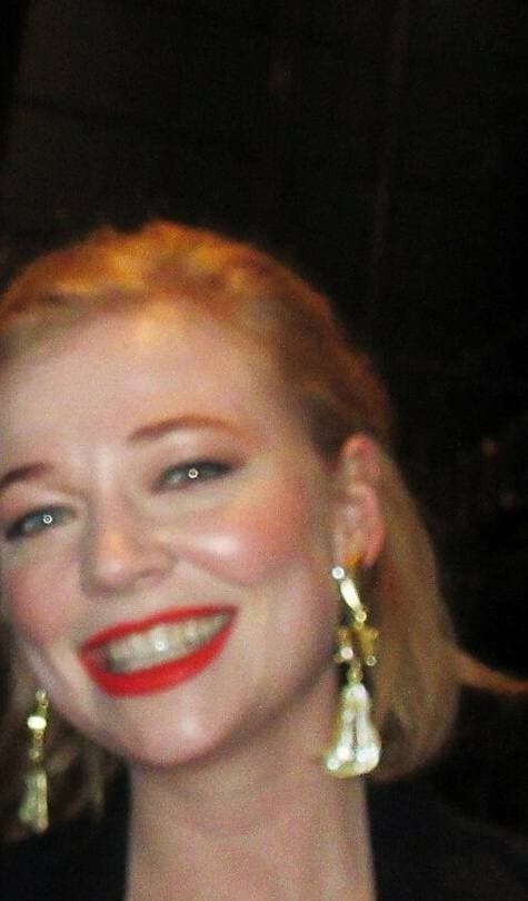 Sarah Snook smiling, red hair pinned back, red lipstick and long gold earrings