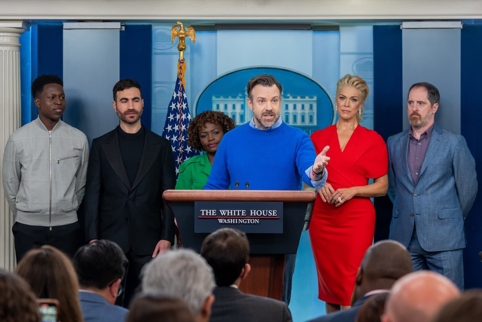 Five members of the Ted Lasso cast standing behind and beside a lectern, Jason Sudeikis speaking in a blue jumper