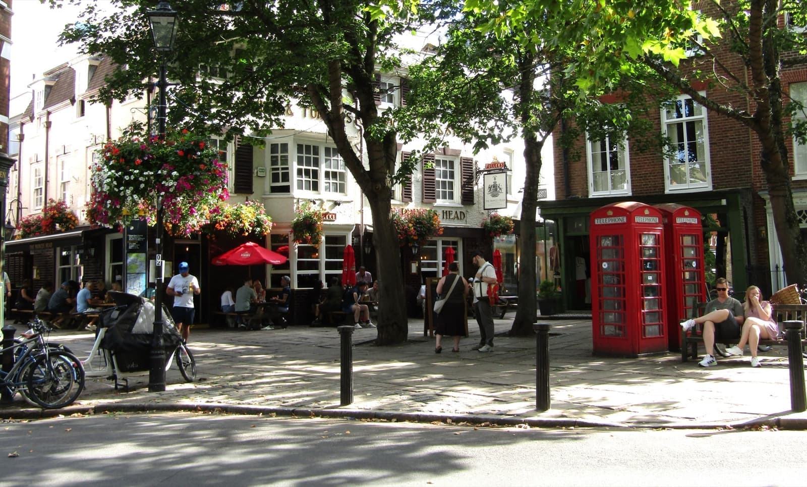 A white-painted corner pub behind trees, with a red telephone box and people sitting outside