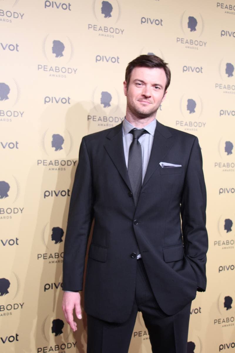 Costa Ronin in a dark suit in front of a sponsor wall