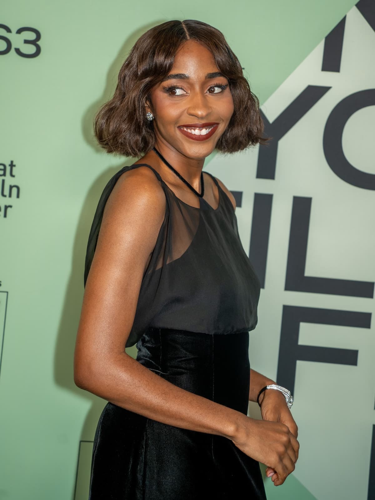 Ayo Edebiri in a black sheer dress, smiling over her shoulder in front of a pale green wall