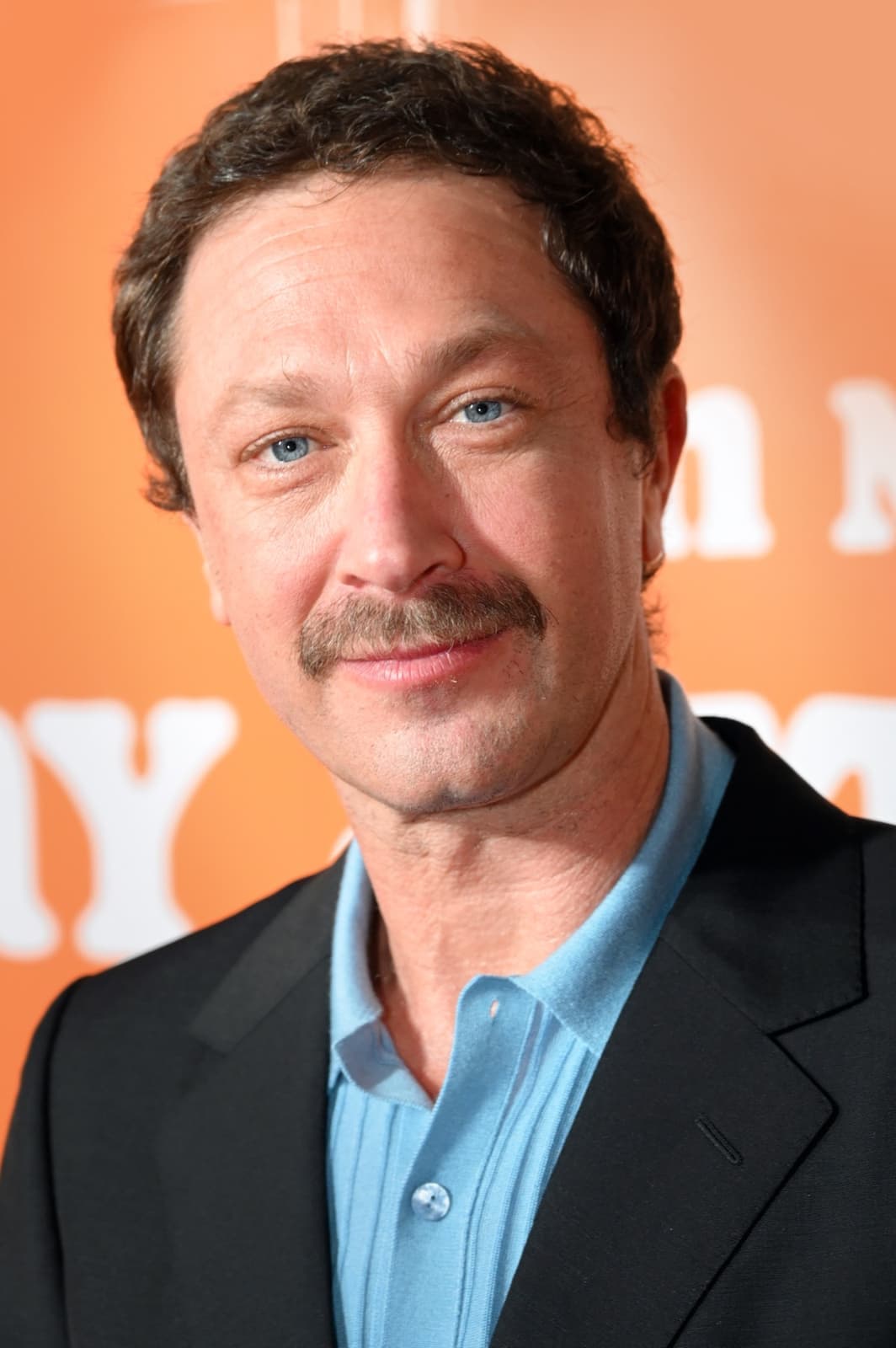 Ebon Moss-Bachrach, moustached, in a dark jacket and blue shirt, smiling against an orange backdrop