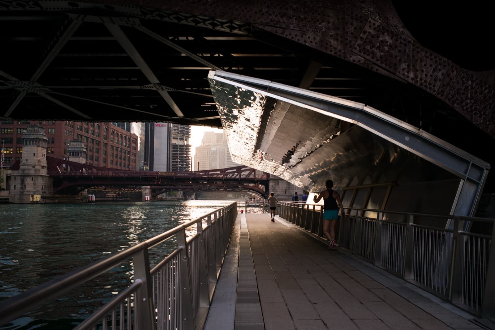 A riverside walkway passing under the dark steel of a bridge, the river bright beyond