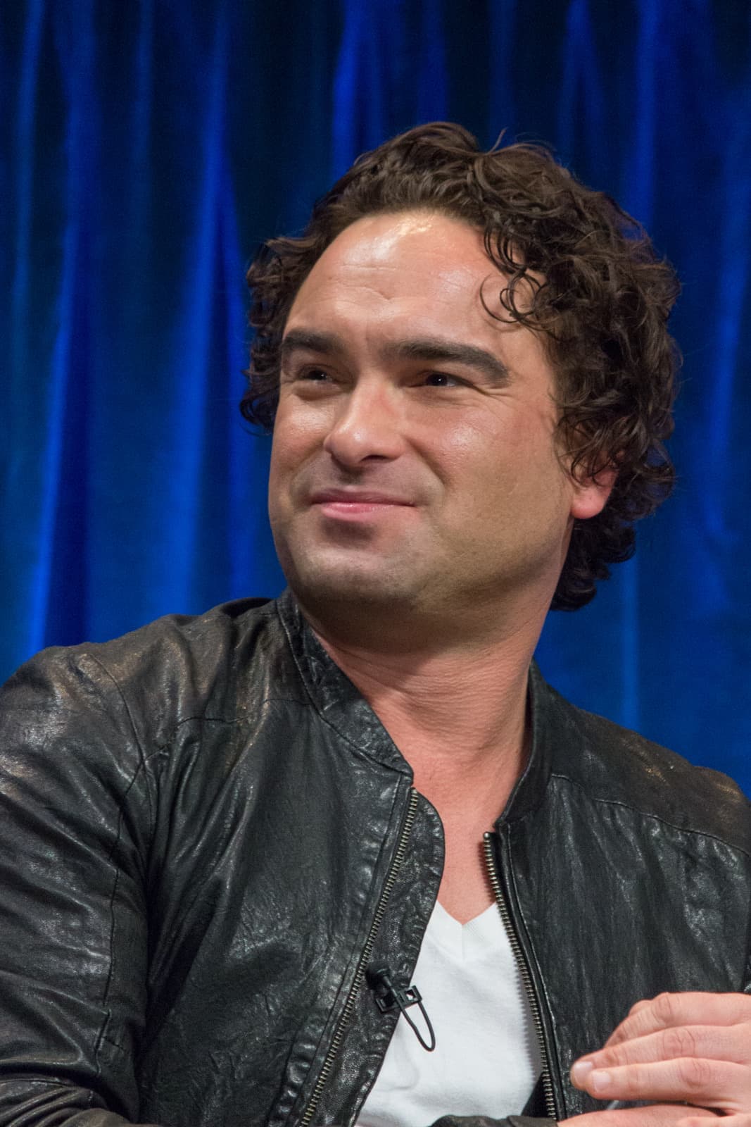Johnny Galecki in a leather jacket, smiling, against a blue curtain