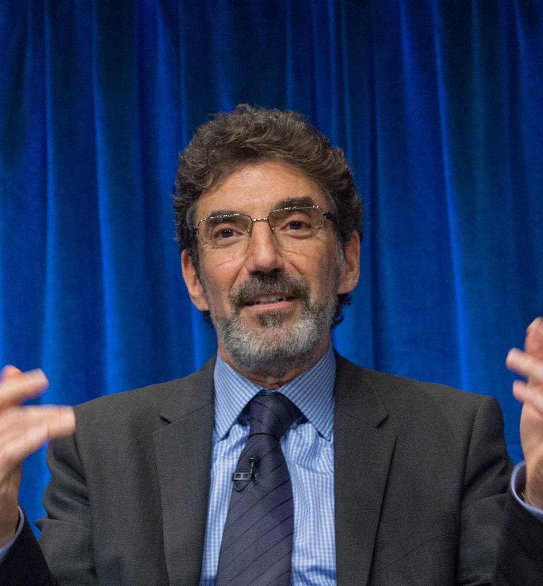 Chuck Lorre, grey-bearded, in a dark suit and blue tie, speaking on stage