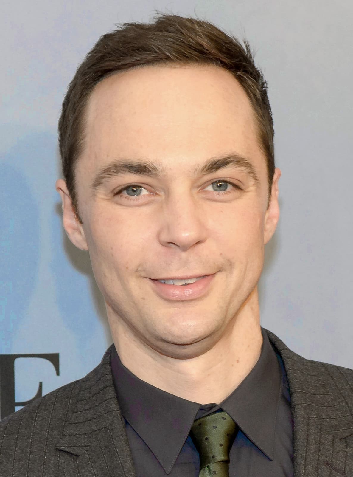 Jim Parsons smiling in a dark suit and green tie against a pale backdrop
