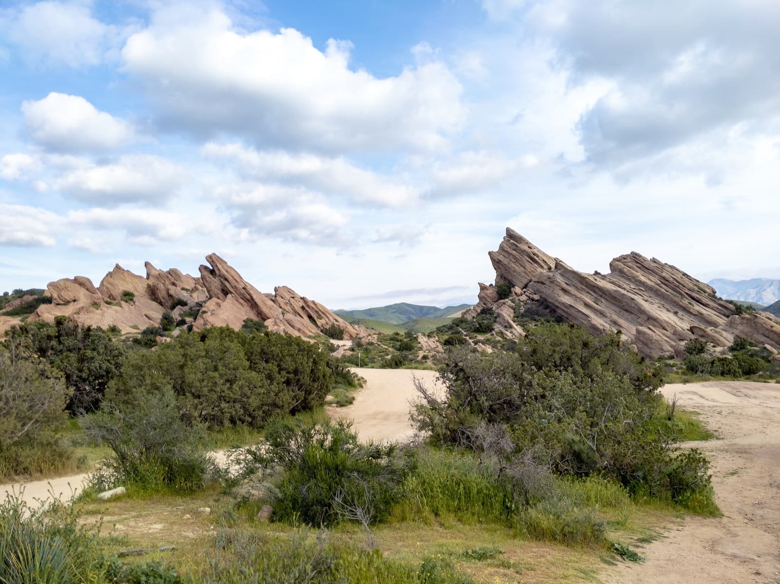 Tilted slabs of pink sandstone rising out of green scrub under a cloudy blue sky