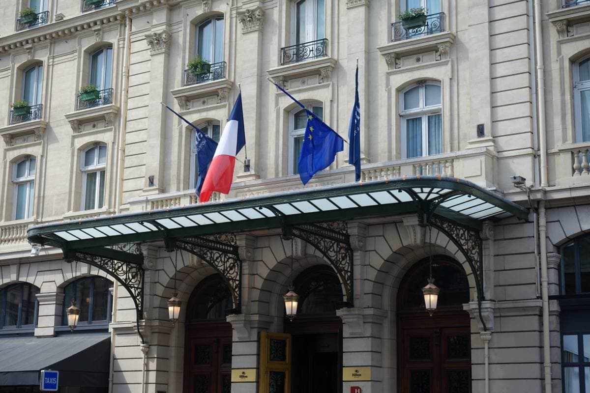 A grand stone hotel front with a glass canopy and flags