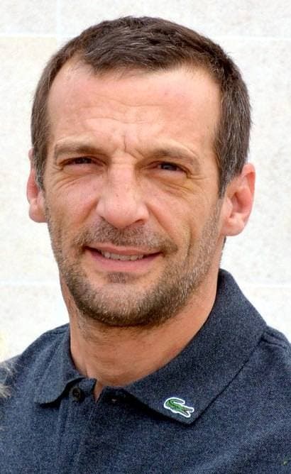 Mathieu Kassovitz, short dark hair and stubble, in a navy polo shirt
