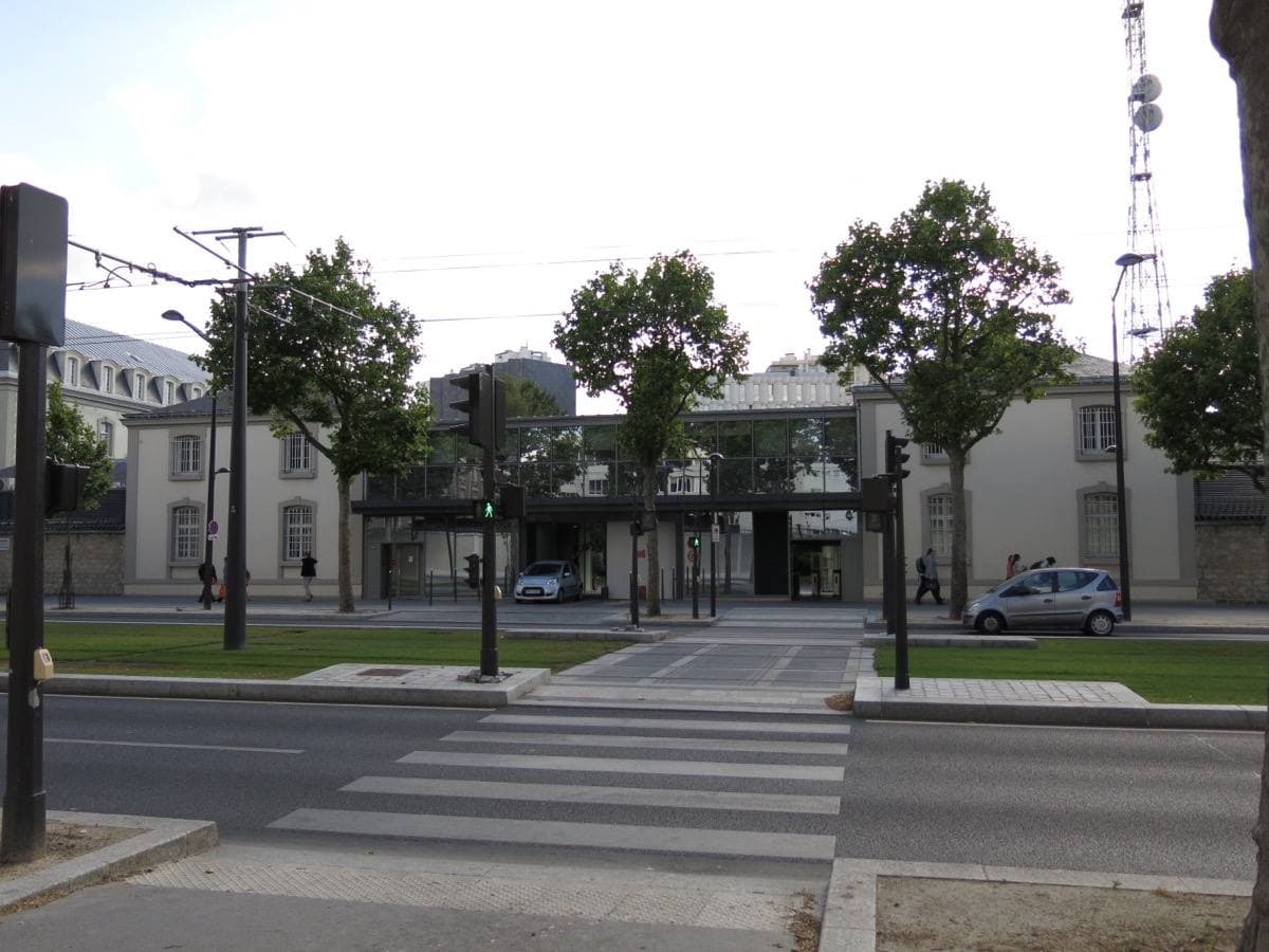 A long low wall and gatehouse behind young trees on a wide boulevard