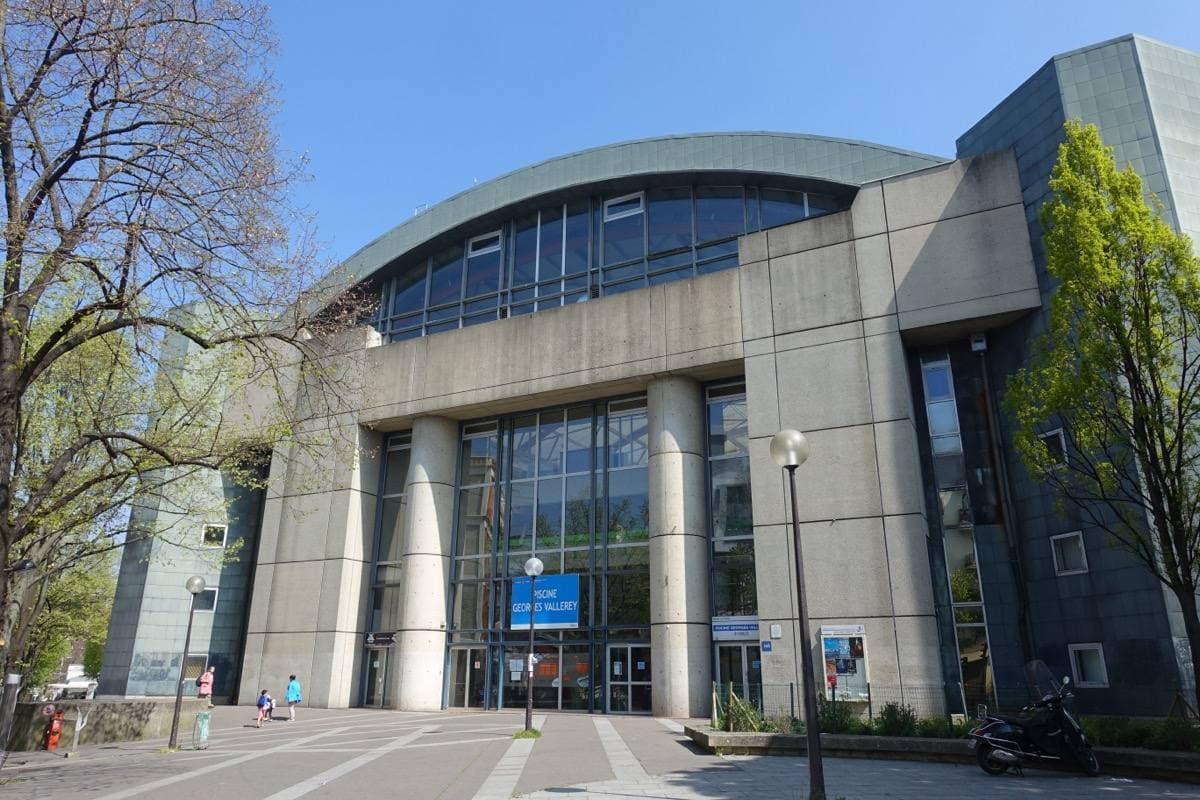 A curved glass and concrete swimming-pool building on a sunny square