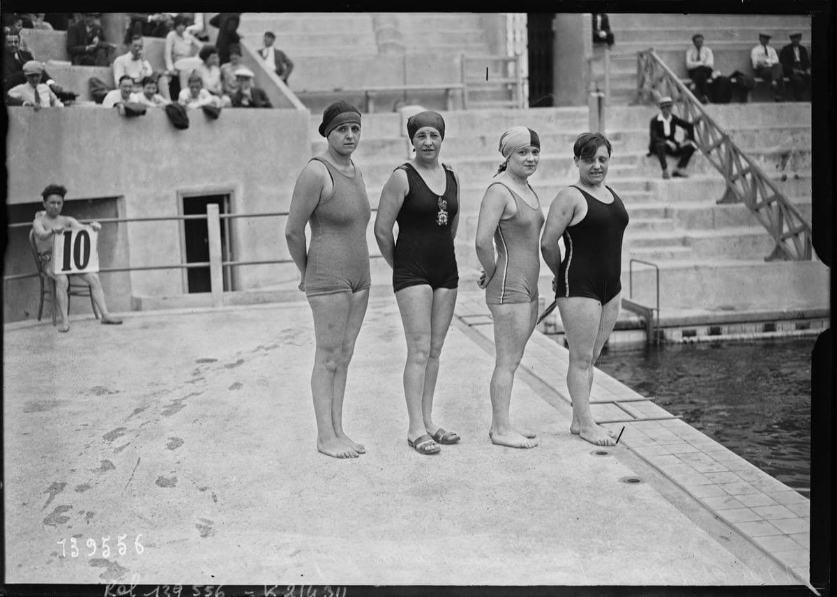 Four women in black swimsuits and caps on a pool deck, a crowd on the stands behind