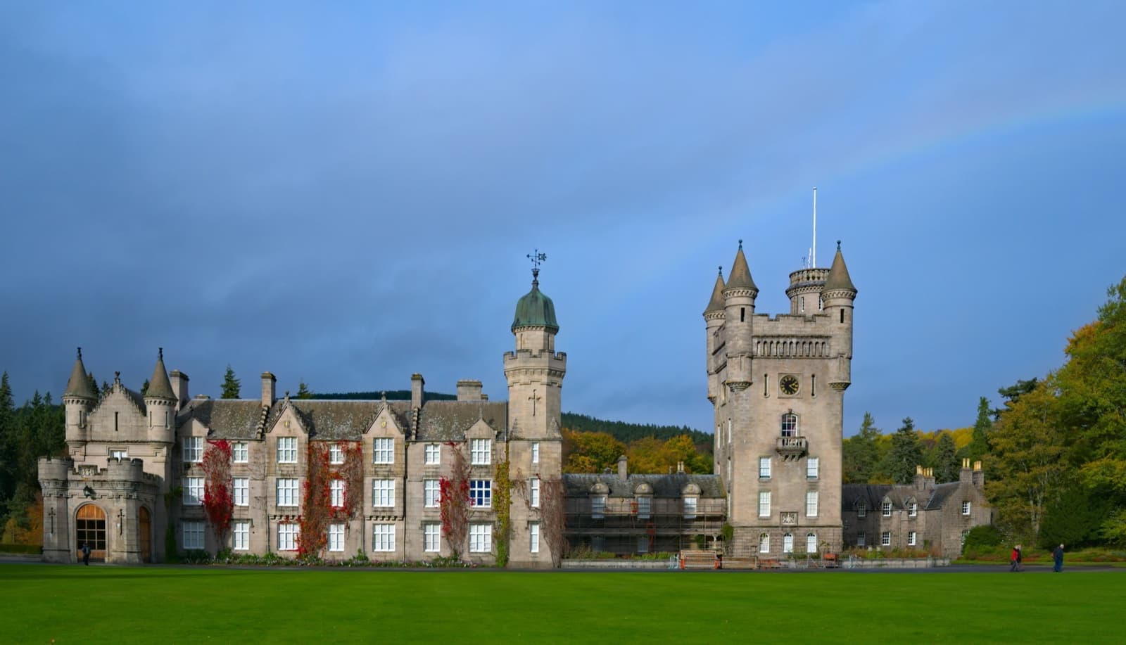 A granite castle with a tall clock tower behind a wide green lawn, under a blue sky