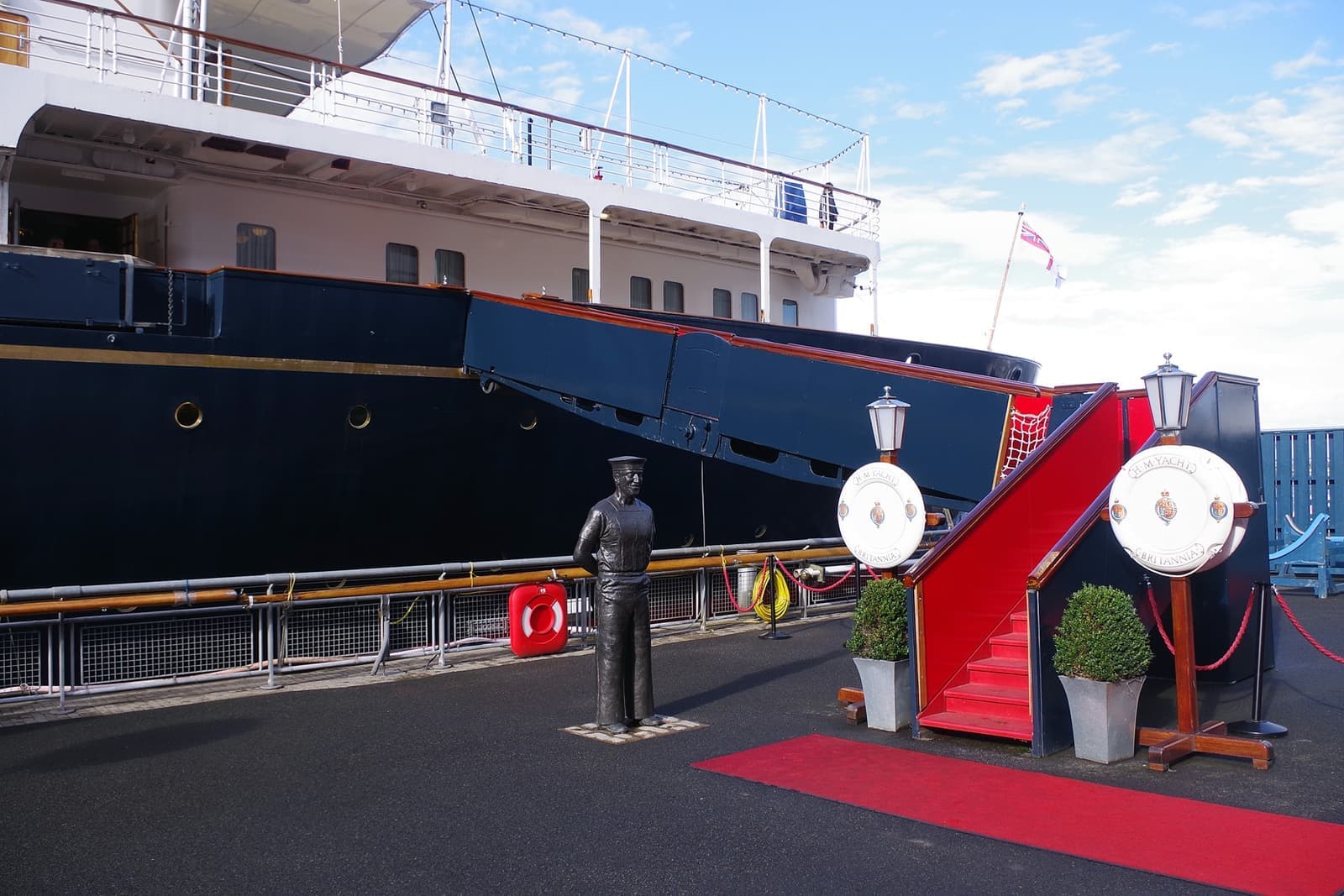 The dark blue hull of the Royal Yacht Britannia beside a quay, with a red-carpeted gangway and a bronze statue