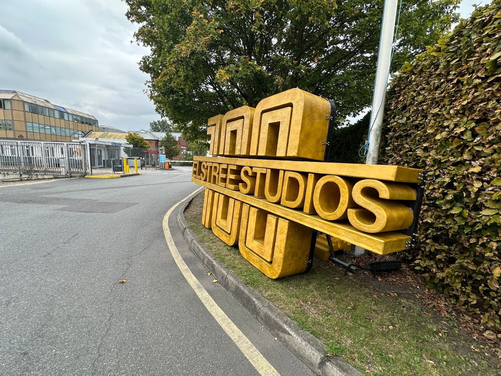 A curving studio road past a large gold sign reading Elstree Studios, with low studio buildings behind