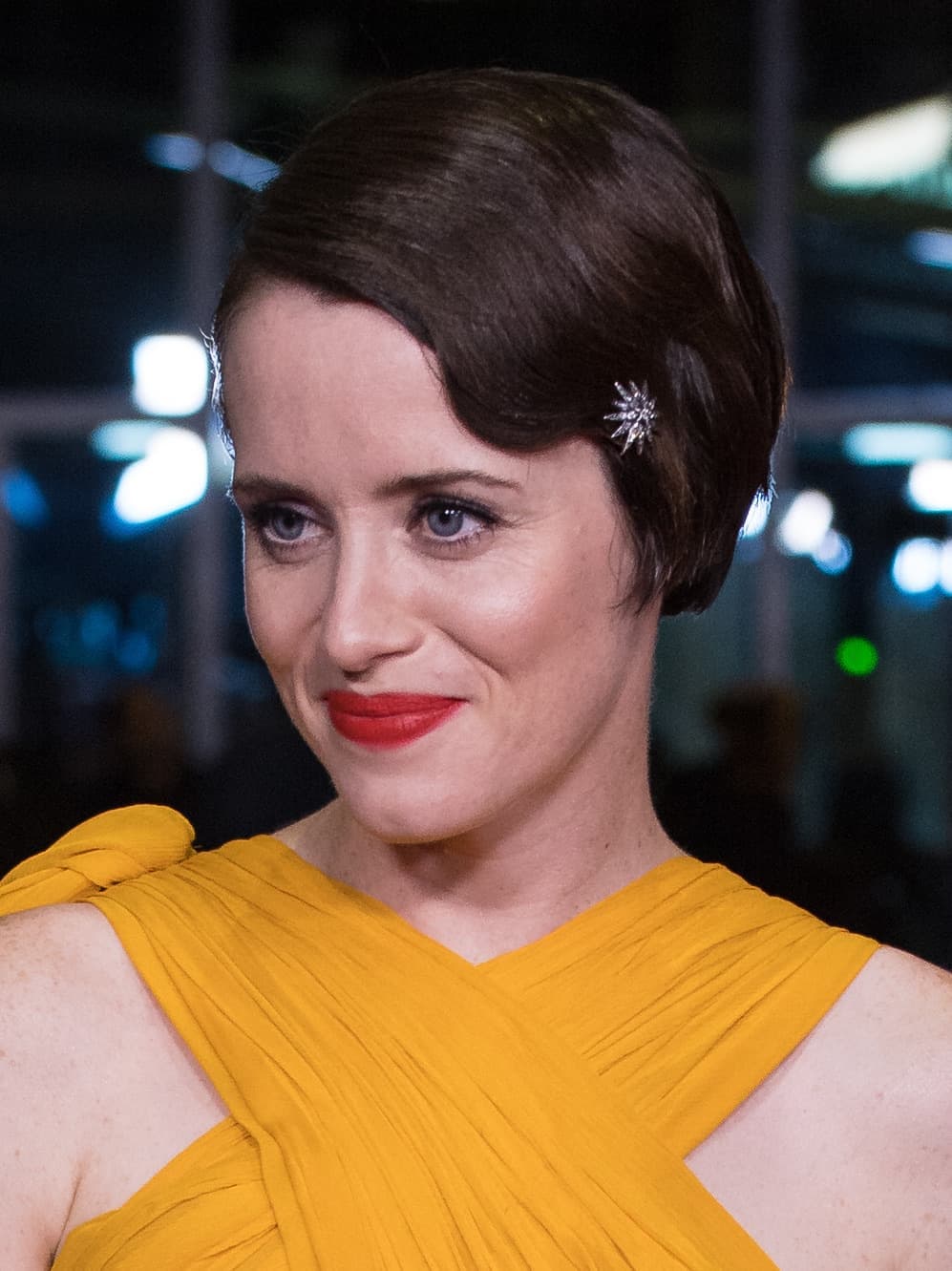 Claire Foy with short dark hair, red lipstick and a mustard-yellow dress, smiling to one side