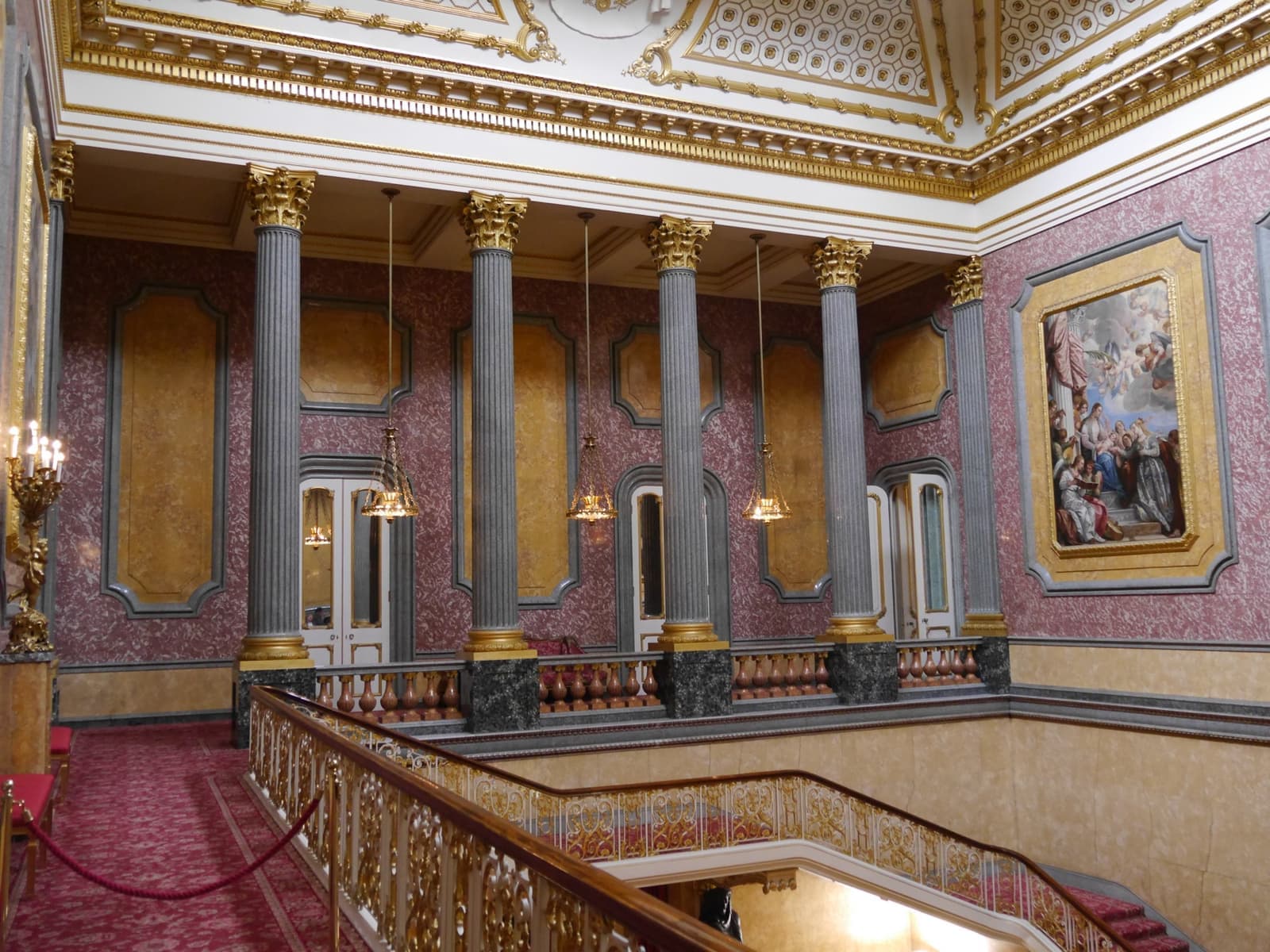 A gilded staircase hall with grey marble columns, a red-carpeted stair, chandeliers and large oil paintings