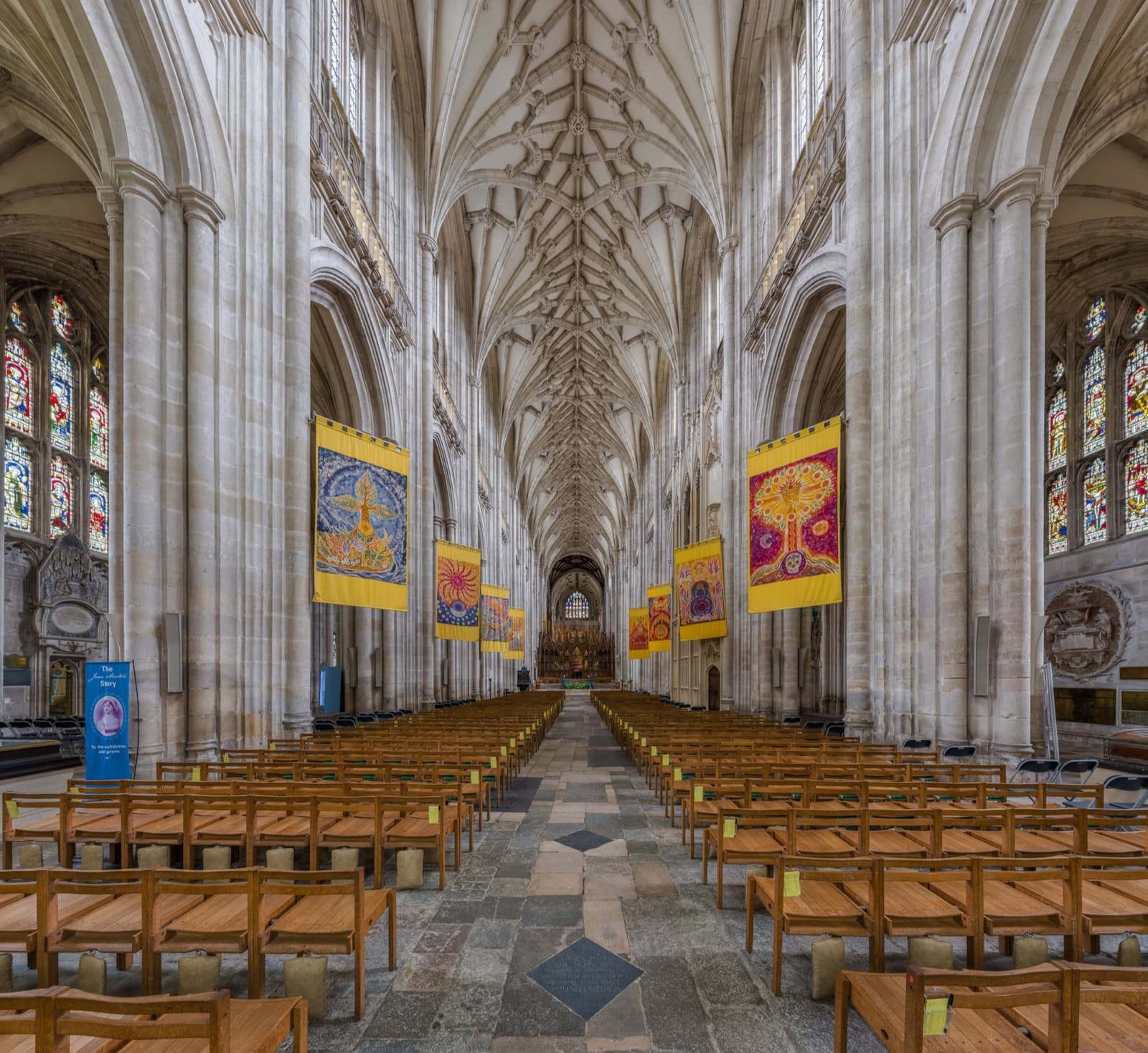A long Gothic nave of pale stone with a vaulted roof, rows of wooden chairs and hanging banners